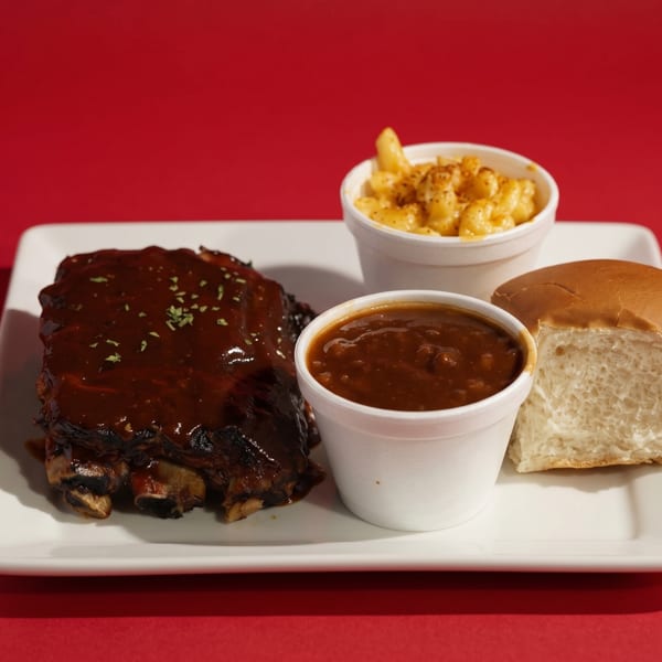BBQ ribs with sauce, baked beans, mac and cheese, and a dinner roll on a white plate.