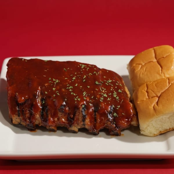 A rack of BBQ ribs with sauce and parsley, served with two dinner rolls on a white plate.