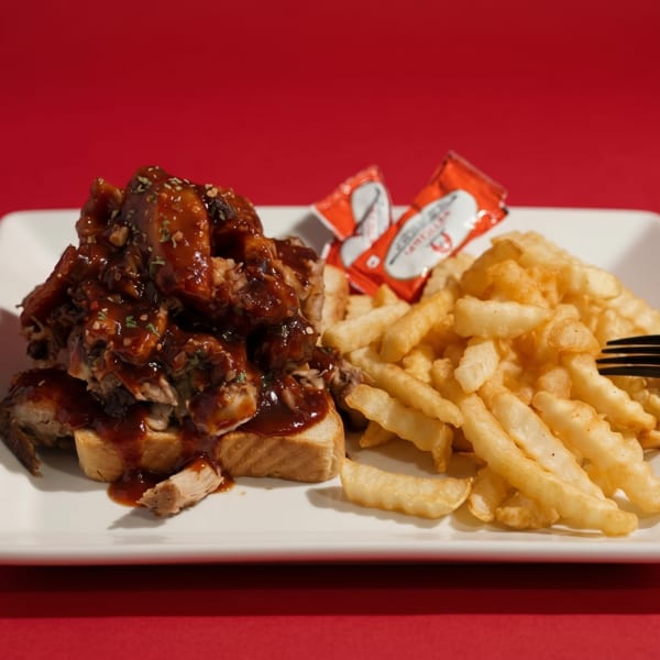 BBQ chicken sandwich with fries and ketchup packets on a white plate against a red background.